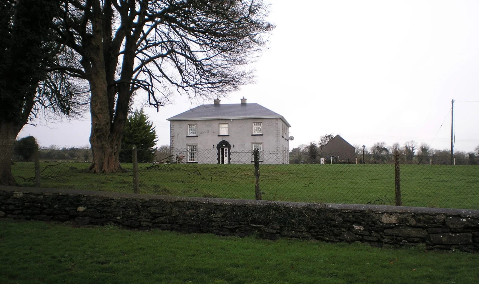 Carrowroe House, a two-storey grey rendered farmhouse behind a stone wall and tall trees