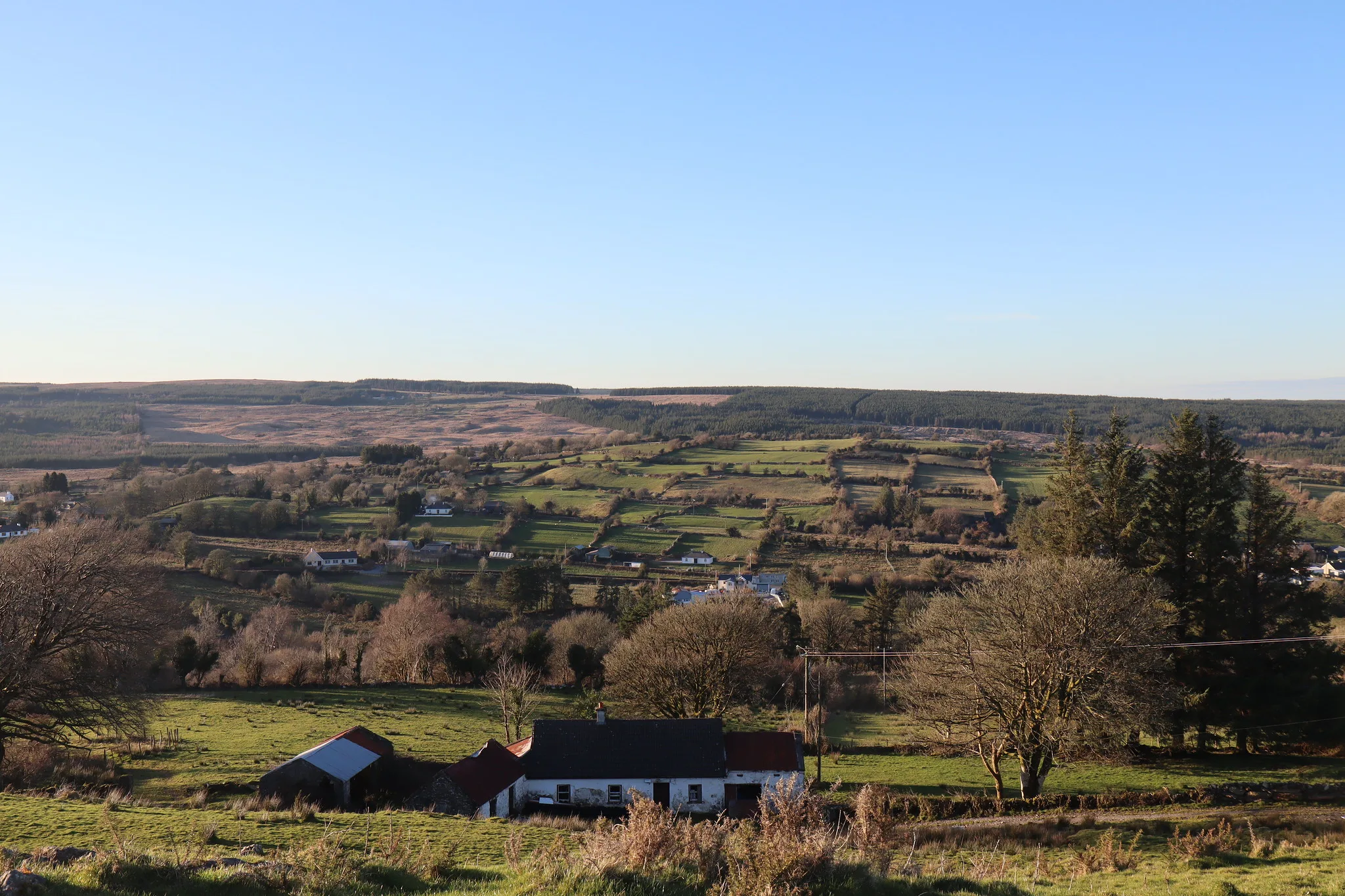 An old farmhouse with a stone outbuilding, seen from a hillside above a wide valley of fields and forestry